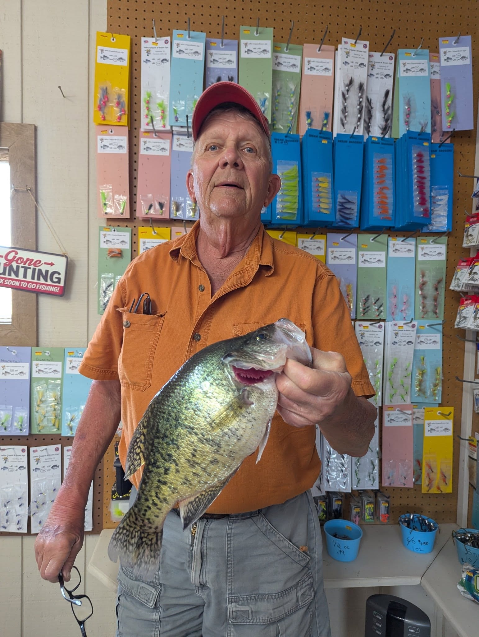 Man in orange shirt holding a large crappie in front of the jig display wall