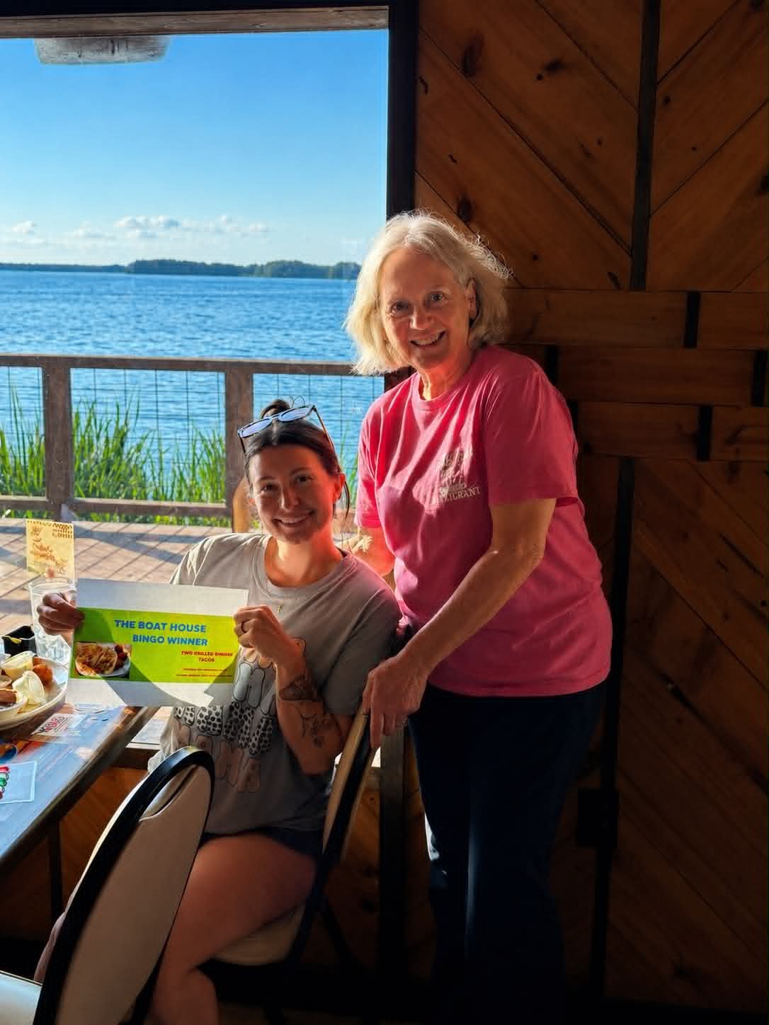 Boat House Bingo winner holding prize certificate with lake view at The Boat House Restaurant