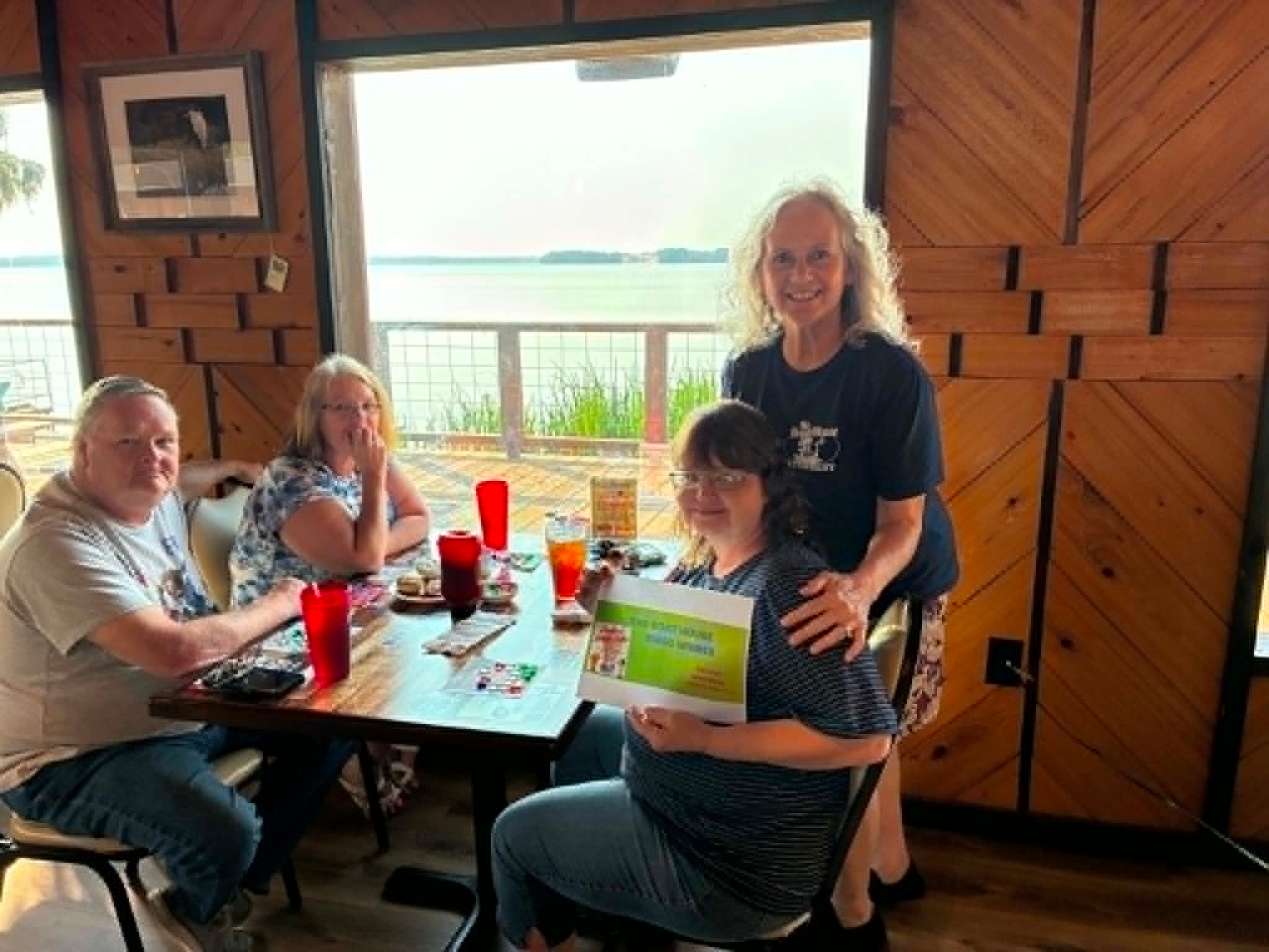 Boat House Bingo winner holding certificate with friends at lakeside table