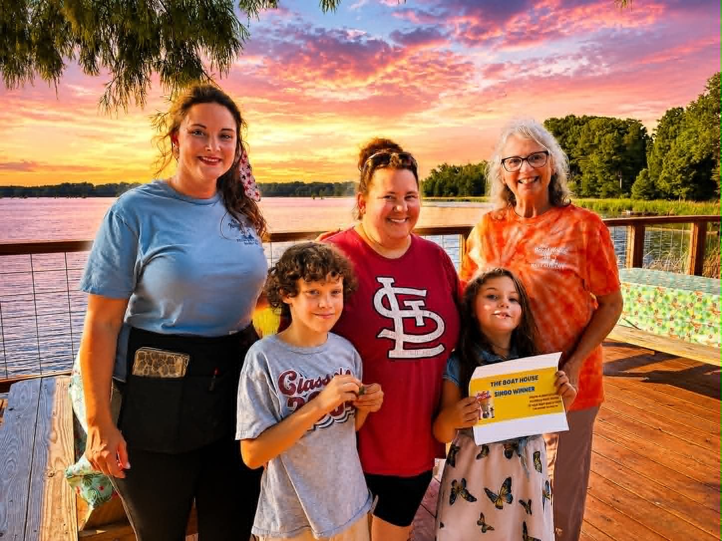 Family of five holding a Boat House Bingo winner certificate on the deck at sunset over Reelfoot Lake