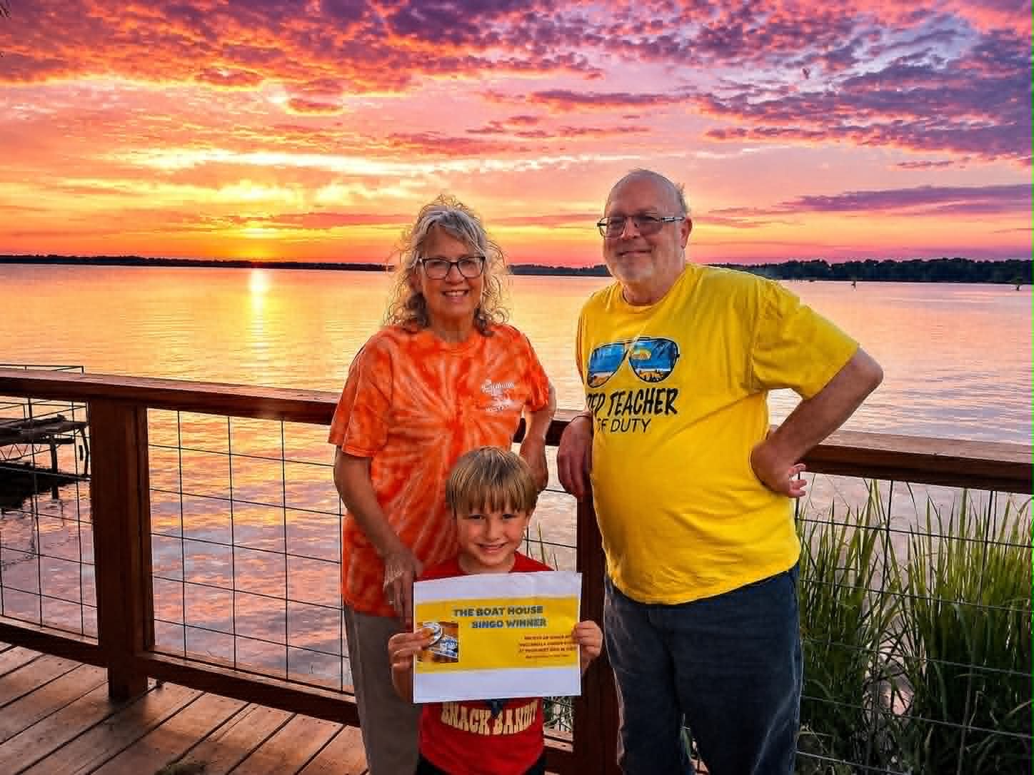 Grandparents and grandson holding a Boat House Bingo winner certificate on the deck at sunset