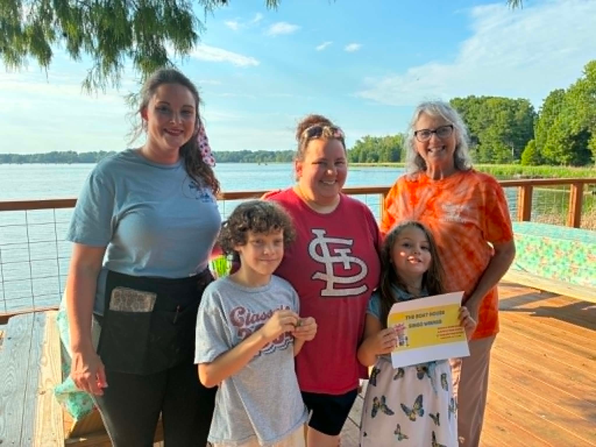 Family of five with Boat House Bingo winner certificate on the lakeside deck in afternoon light